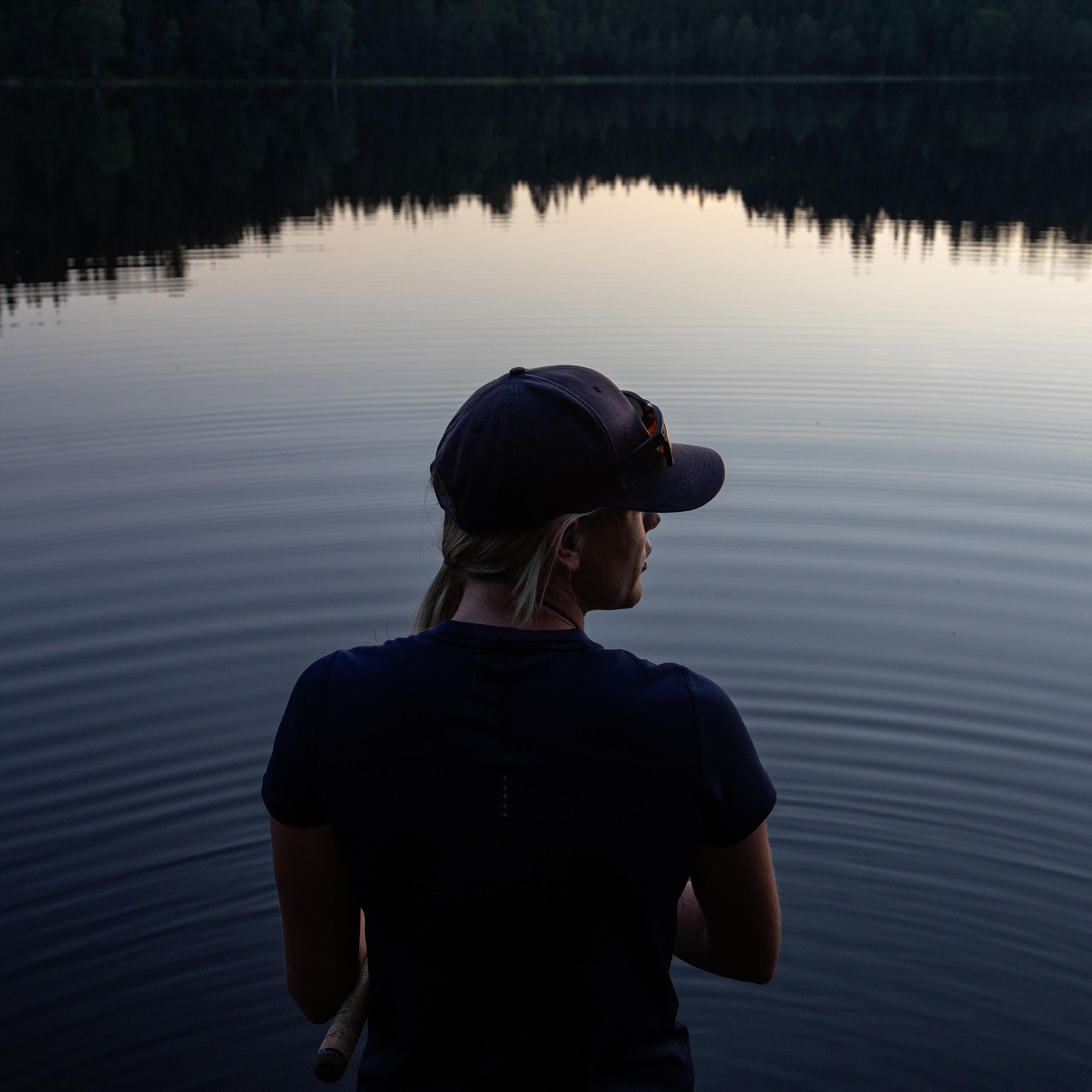 Nærbilde av silhuetten til en kvinne som står med ryggen til i blå t-skjorte og caps og fisker i skumringen. 