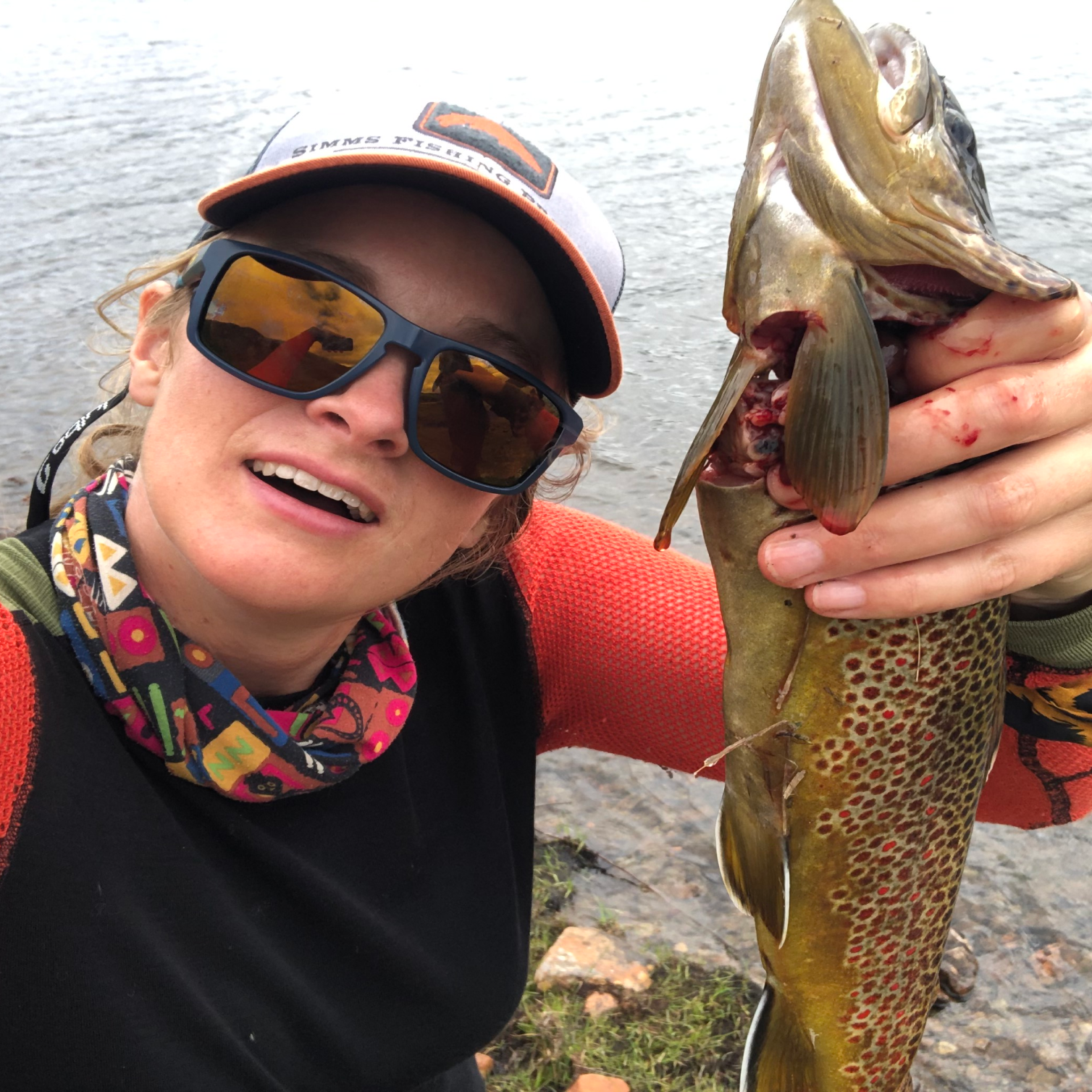Bilde av Inger Marie Berling i Fiskejenter i Marka tatt ovenfra og ned som en selfie. Hun holder en stor nyfisket ørret i den ene hånden mens hun kikker inn i kameraet. Hun har på seg caps,  solbriller og ulltrøye i rødt og sort.