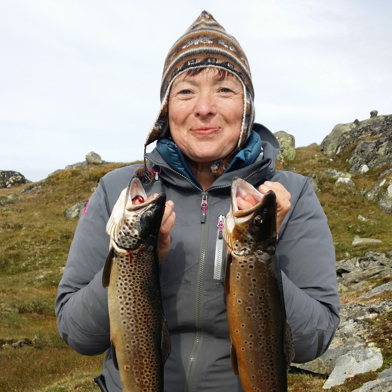 Bilde av Kristin Fjellestad i Fiskejenter i Marka tatt fra livet og opp i et fjellandskap. Hun smiler lurt og holder en stor ørret i hver hånd. Fiskene henger rett ned. Hun har på seg brun og hvit topplue og grå skalljakke.