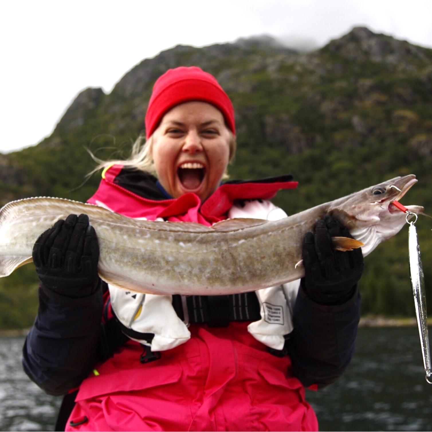 Bilde av Marthe Louise Rossing Fosser i Fiskejenter i Marka som smiler euforisk mens hun holder en stor lange i hendene foran seg. Fisken har en lang sluk hengende ut av munnviken. Marthe har blondt mellomlangt hår og har på seg rosa lue og jakke og hvit redningsvest.