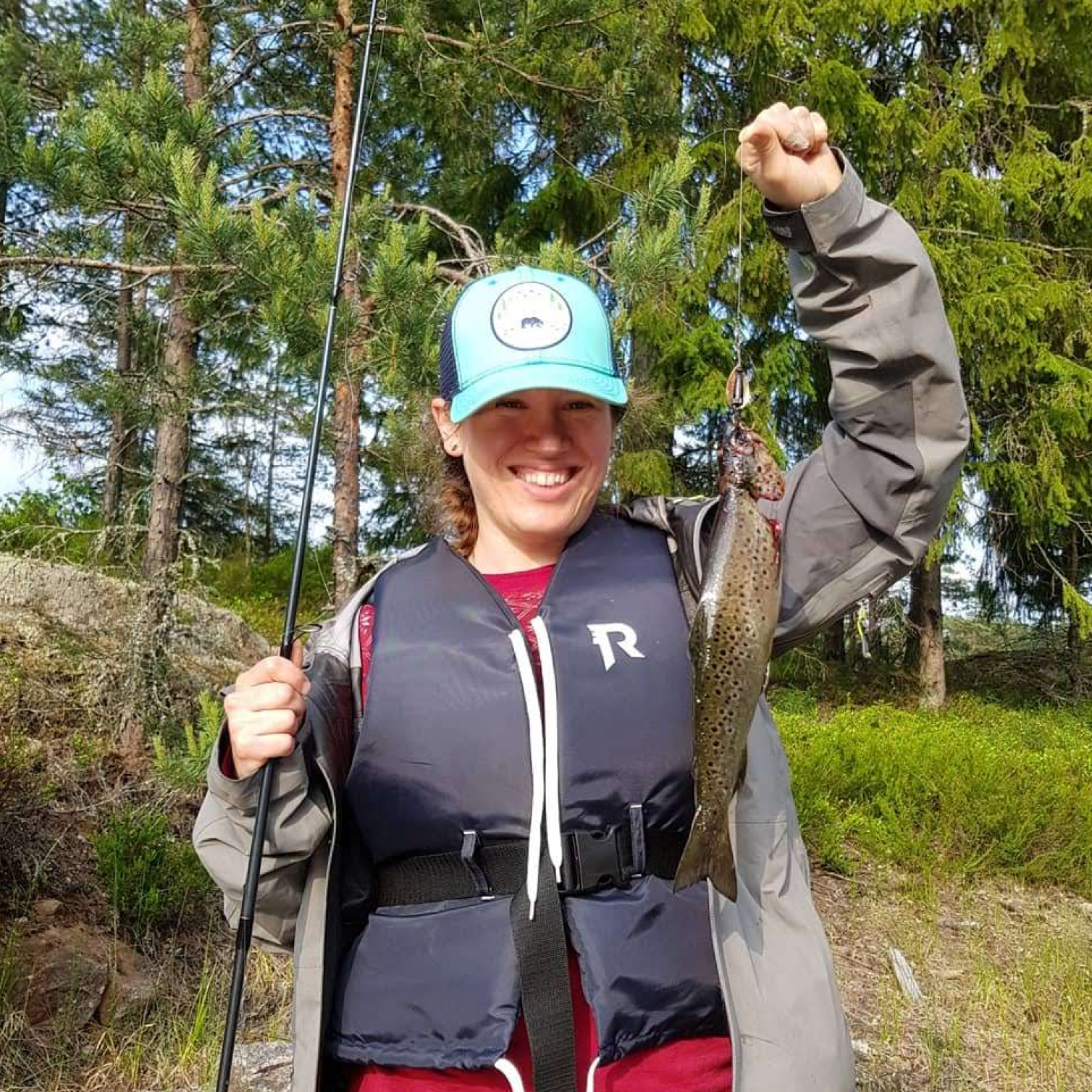 Bilde av en smilende Signe Alexandra Domogalla i Fiskejenter i Marka fra livet og opp. Hun står og holder en fiskestang i ene hånden og en ørret som henger fra en sluk i andre hånden. Hun har på seg caps, redningsvest og friluftsklær. Håret er brunt og flettet.