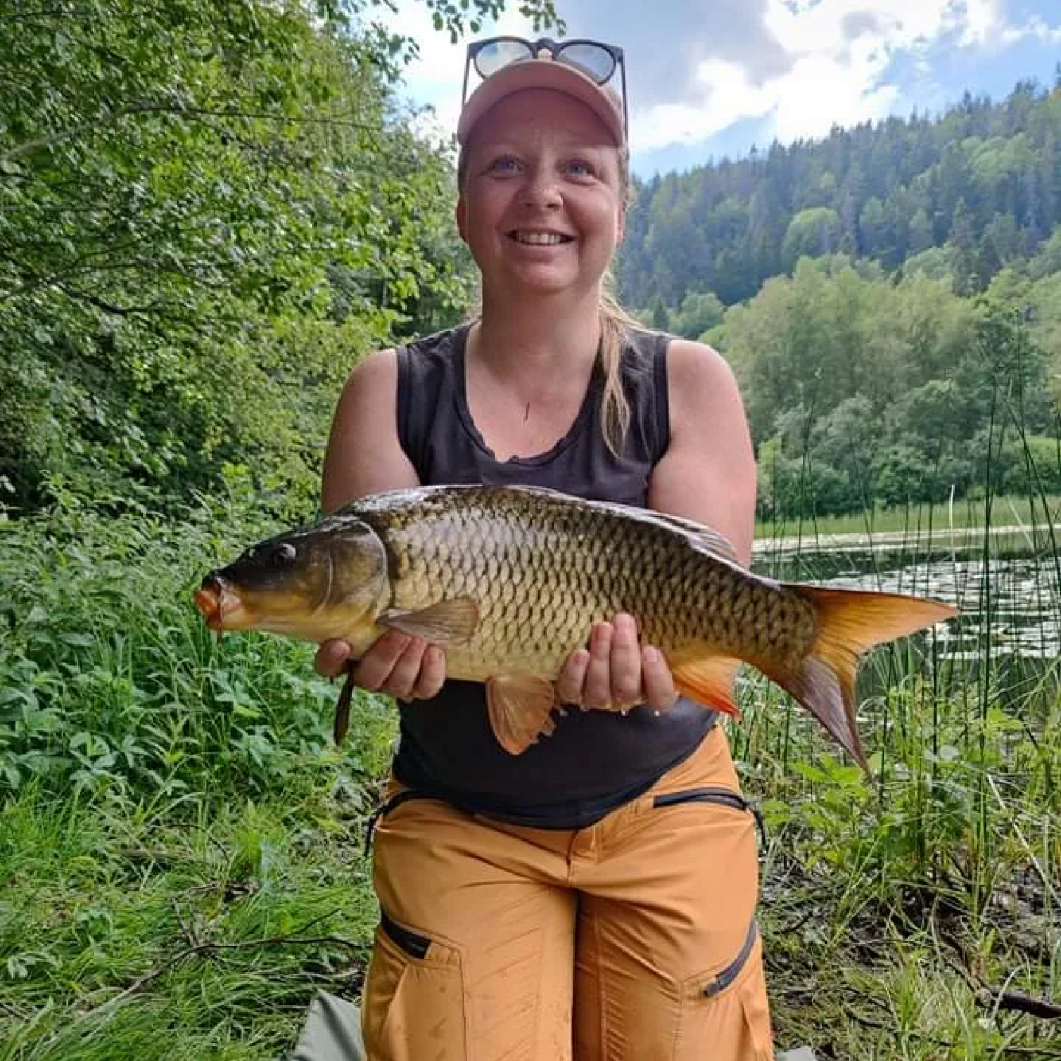 Bilde av en smilende Synnøve Moss i Fiskejenter i Marka som sitter på knærne ved et vann mens hun holder en karpe i hendene foran seg. Hun har langt blondt hår i hestehale og har på seg caps med solbriller på hodet. Hun har svart singlet og orange bukser.